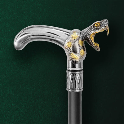 Close-up of a Black Fang Walking Cane handle featuring a detailed silver snake design with gold accents and fangs wrapped around the curved grip against a dark green background.