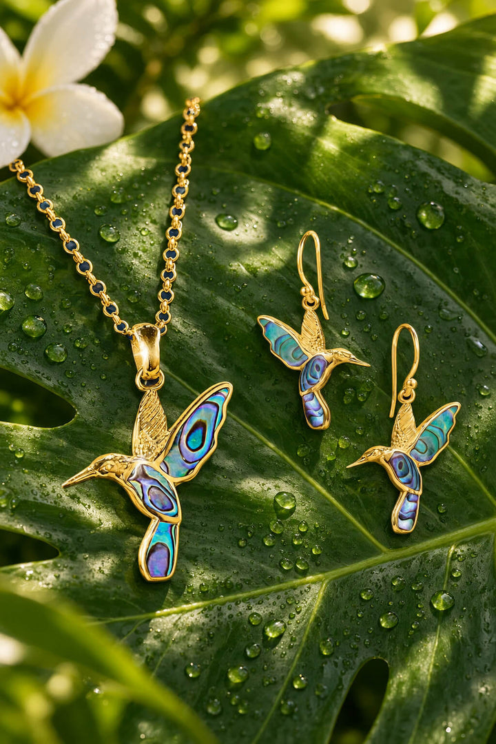 Golden Abalone Hummingbird Collection featuring a gold tone hummingbird necklace and matching earrings with blue abalone accents on a dewy tropical leaf Up To 75% Off Retail