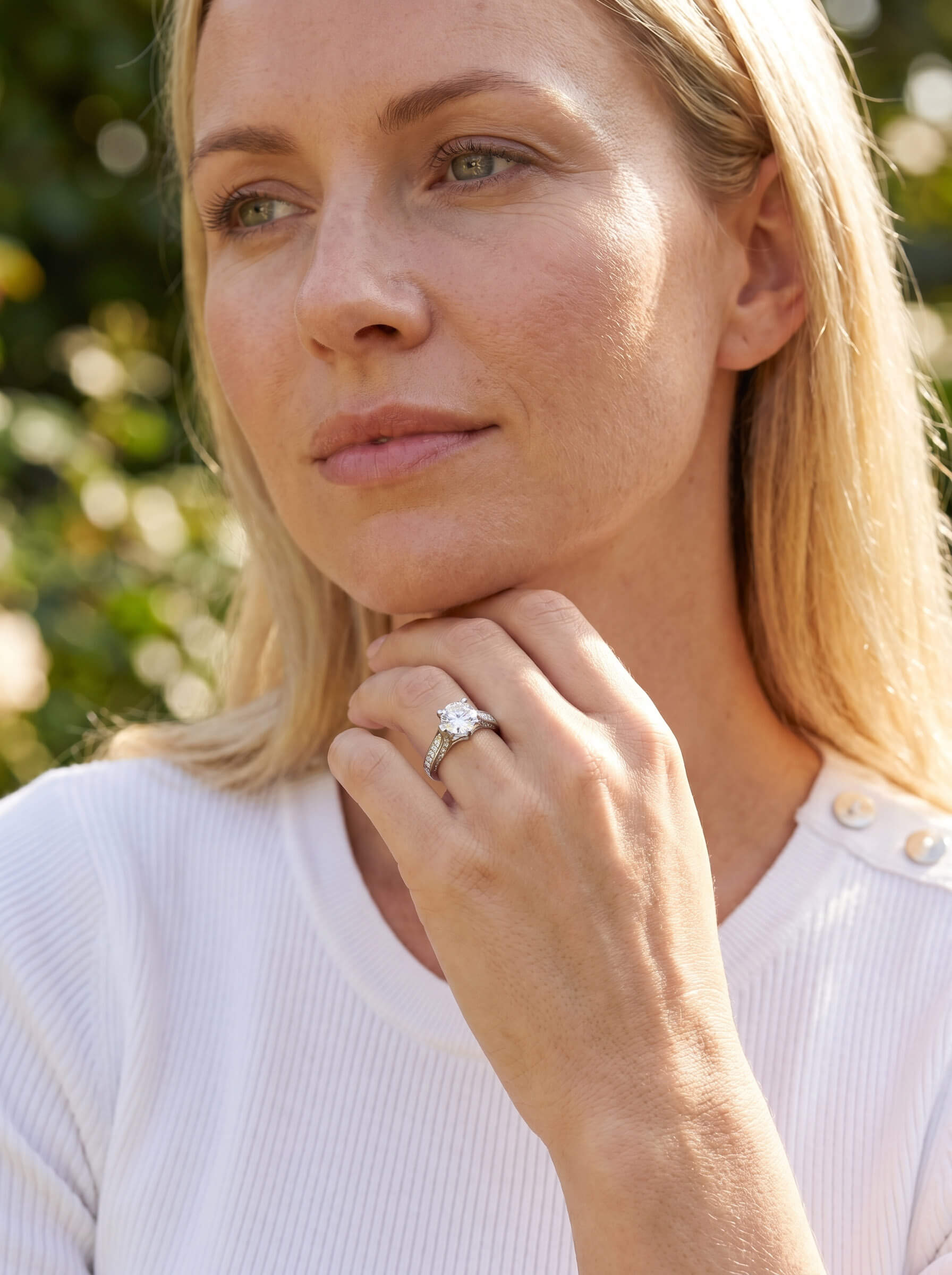 Woman wearing a Novalyn Moissanite Ring with a large center stone and sparkling band, posing thoughtfully outdoors in natural light.
