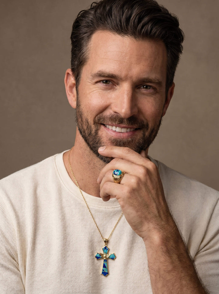 Man wearing a Wings Of Freedom Ring with a blue stone, smiling and resting his hand near his face, also wearing a gold necklace with a blue cross pendant.