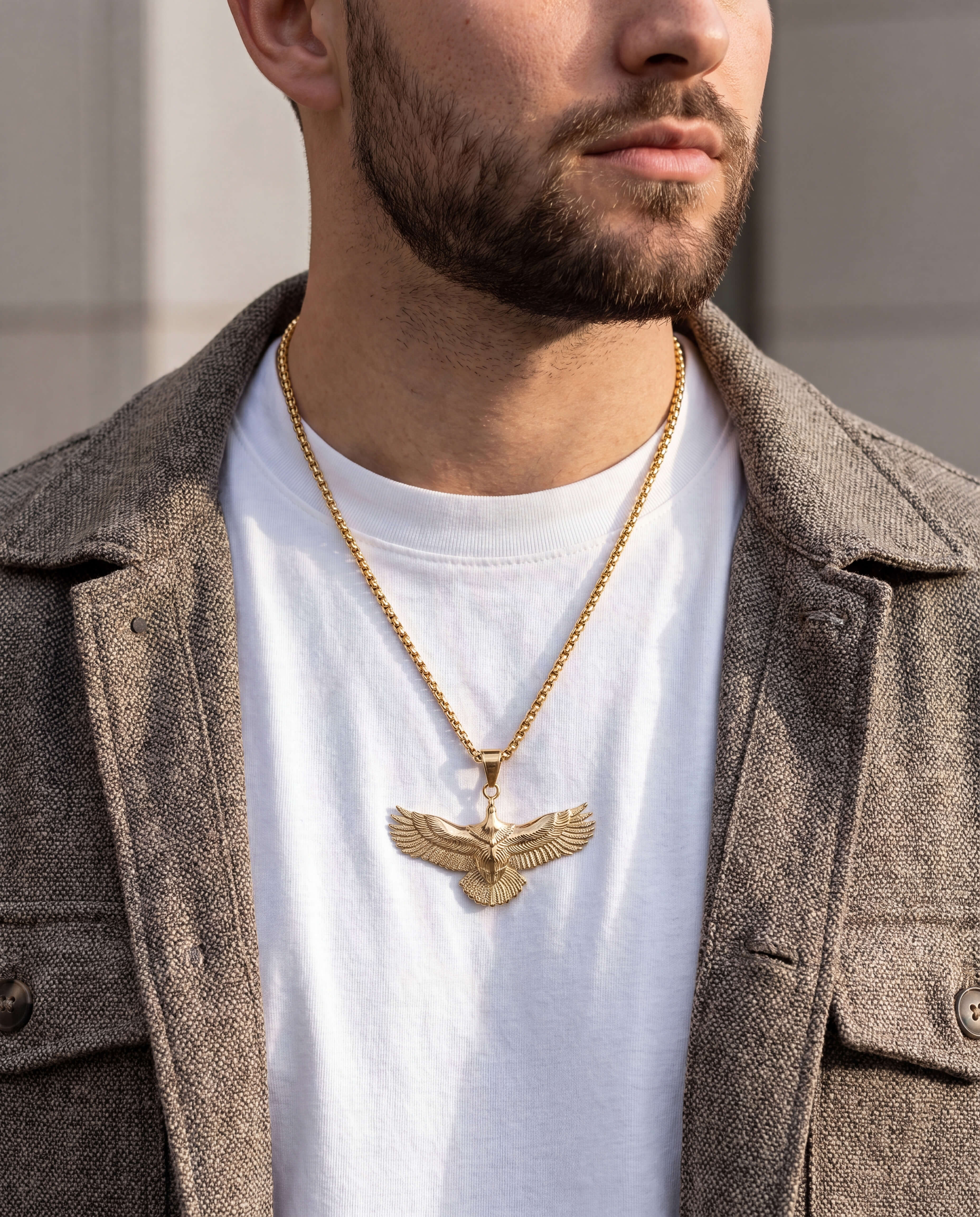 Man wearing a gold necklace with an eagle pendant, dressed in a white shirt and brown coat.