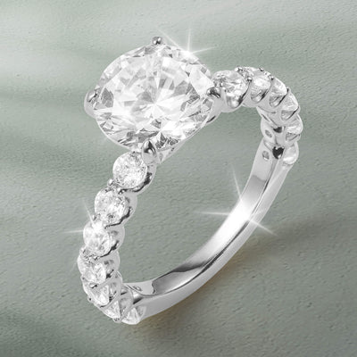 Grandeur Radiance Ring featuring a large faceted center gemstone and a sparkling row of round stones along the polished band on a soft gray background