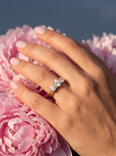 Hand wearing a gold ring with clustered clear stones resting among soft pink peony flowers