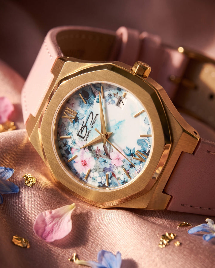 Fleur De Vie Ladies Watch with a gold tone case, floral dial, Roman numerals, and pink leather strap, styled with scattered petals. Up To 75% Off Retail
