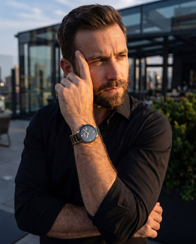 Man wearing a black shirt and watch, standing outdoors with a cityscape background