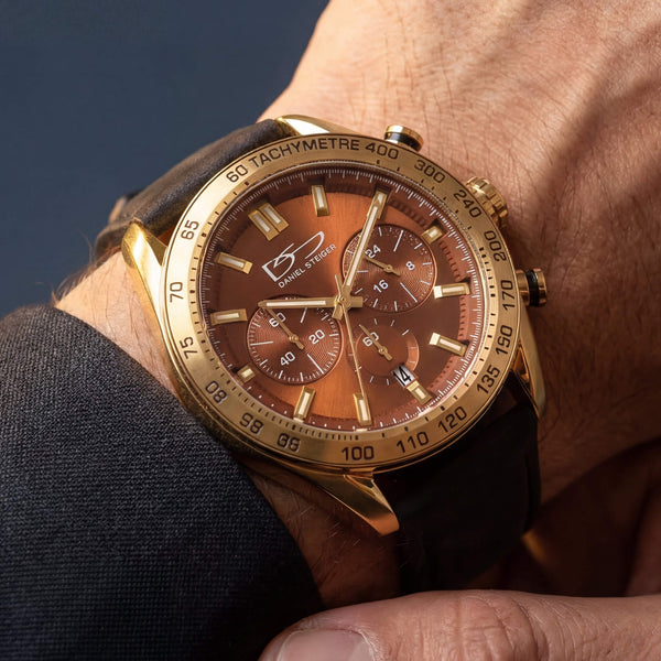 A close-up of a wrist wearing a gold-tone chronograph watch with a brown dial and black leather strap. The watch has a gold bezel with a tachymeter scale, three subdials, and a visible logo on the dial.